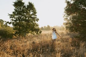 A person in a blue dress stands in a golden field at sunset with tall trees silhouetted in the background.