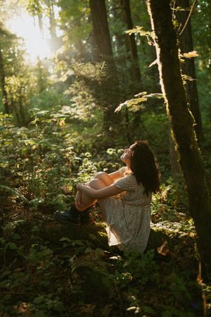 A dreamy forest scene with sunlight filtering through trees as someone sits peacefully on the ground in a white dress.