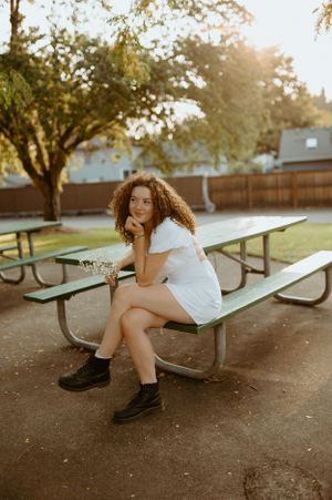 Person in white dress sitting on green park bench at sunset with curly hair and black boots.