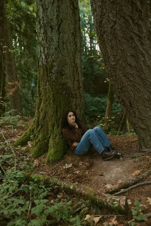 Person sits against mossy tree trunk in lush forest setting with dappled sunlight filtering through canopy.