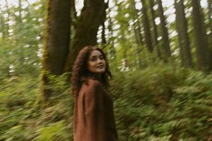 A person in a brown coat walks through a lush green forest with sunlight filtering through the trees.