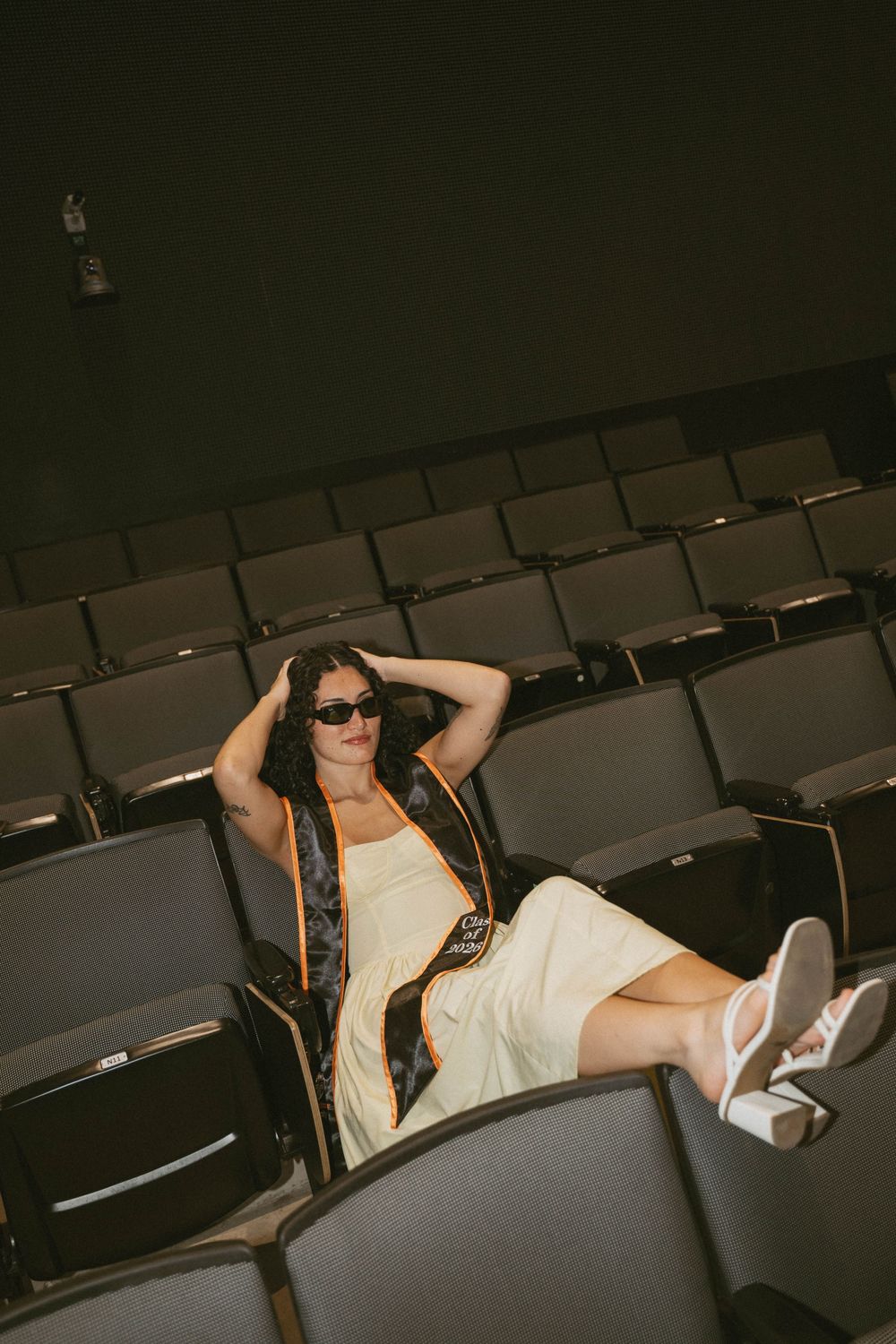 Woman in sunglasses and white dress relaxing in empty theater seats wearing white sneakers and a dark vest.