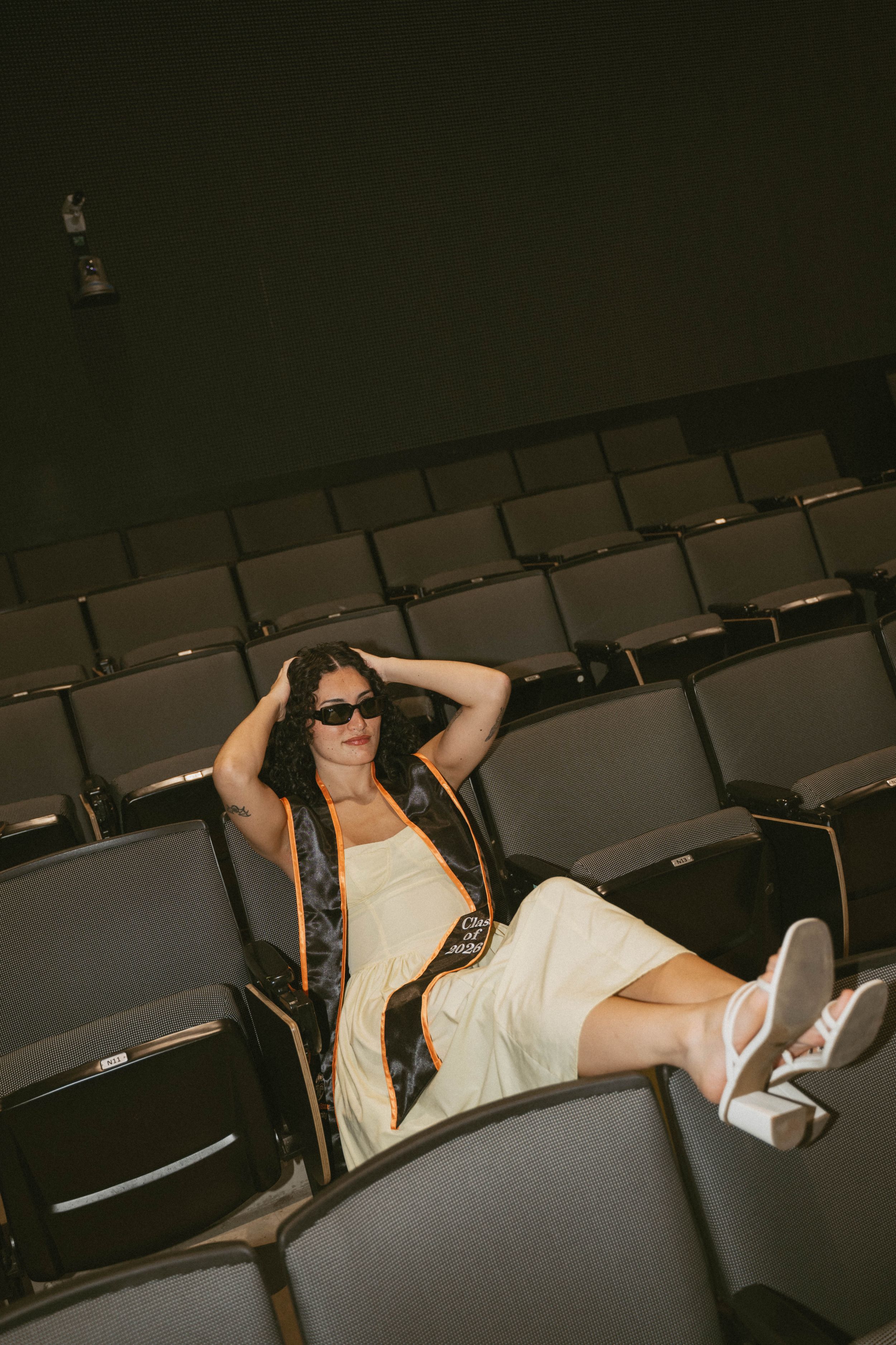 Woman in sunglasses and white dress relaxing in empty theater seats wearing white sneakers and a dark vest.