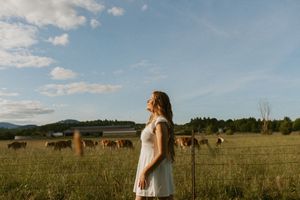 Person in white dress standing in golden field with cattle grazing under blue sky and clouds.