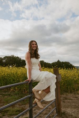 A bride in a white dress sits on a rustic metal gate overlooking a field of yellow wildflowers under cloudy skies.