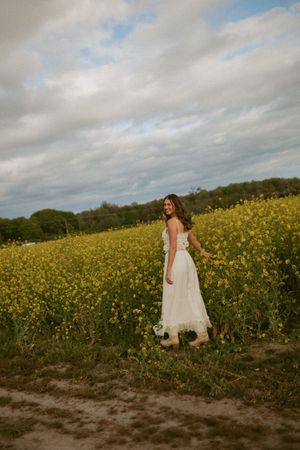 A woman in a white dress stands in a field of yellow wildflowers at golden hour with cloudy skies overhead.