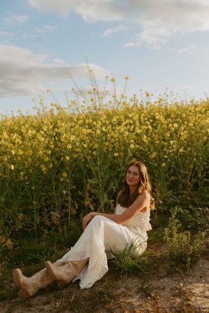 Woman in white dress sitting beside blooming yellow canola field under cloudy sky at sunset.