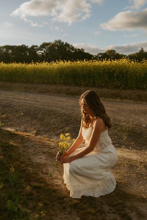 A woman in a flowing white dress kneels on a dirt path beside a vibrant field of yellow flowers at sunset.