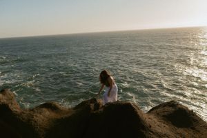 Someone sits on rocky cliffs overlooking the sparkling ocean waves during golden hour sunset.