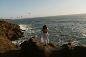 A silhouetted figure stands on rugged coastal cliffs overlooking the Pacific Ocean as seabirds soar in the golden sky.