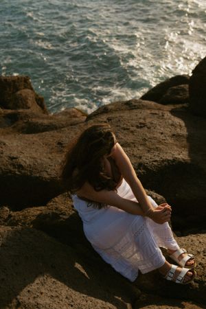 Person in white dress sits on rocky coastal cliff overlooking choppy ocean waters at sunset.