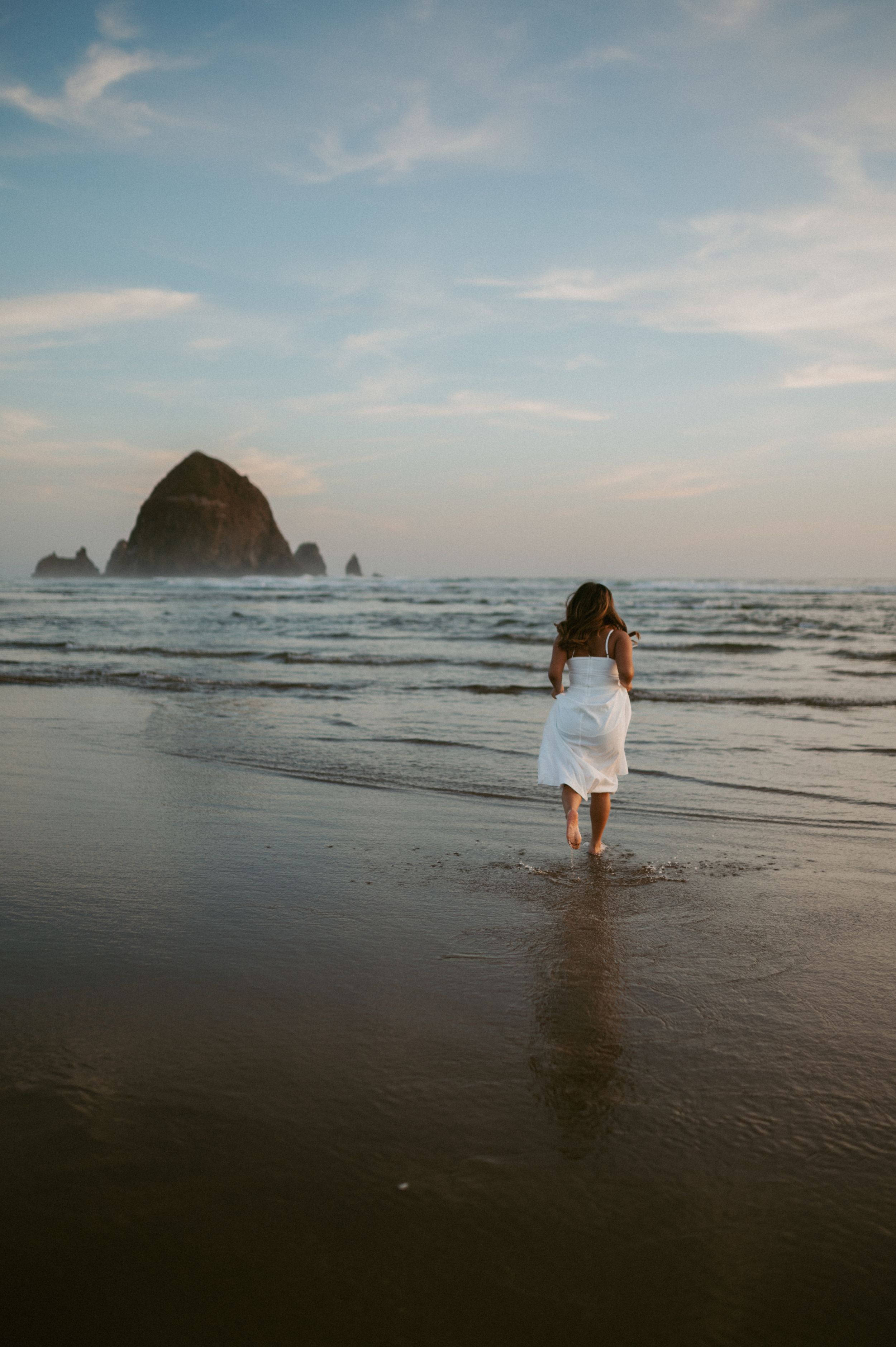 A person in white dress walks along beach near Haystack Rock at sunset in Cannon Beach, Oregon.