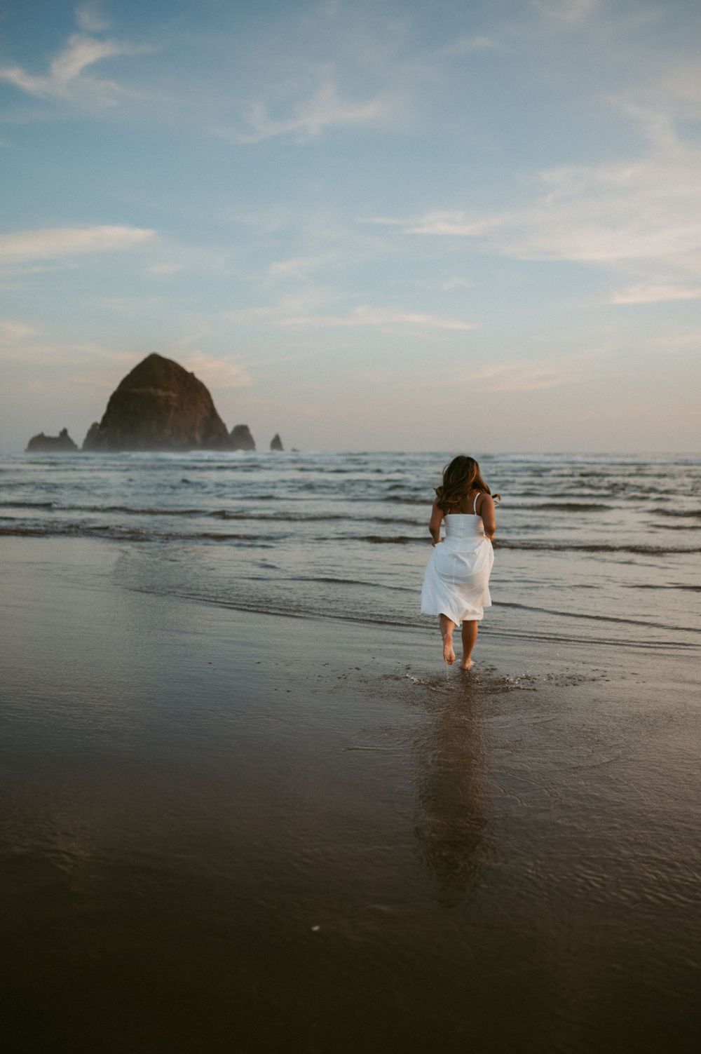 A person in white dress walks along beach near Haystack Rock at sunset in Cannon Beach, Oregon.