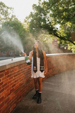 A graduate in white dress and graduation stole sprays champagne while celebrating on a brick walkway at sunset.