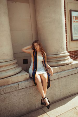 Young woman in white dress and graduation stole poses on stone steps beside classical columns.