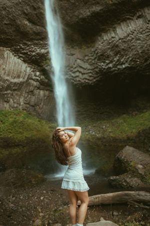 A figure in a white dress poses elegantly near a towering waterfall cascading down a rocky cliff face.