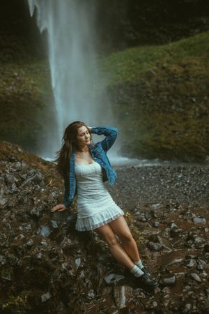 A dreamy portrait scene near a cascading waterfall with rocky terrain and a denim jacket against a moody forest backdrop.
