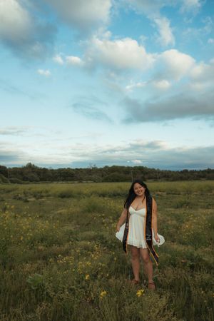 Young woman in white dress holding graduation cap and gown in a green meadow with wildflowers under a cloudy sky.