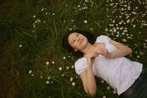 Person lying in green grass surrounded by small white daisies wearing a white blouse on a sunny day.