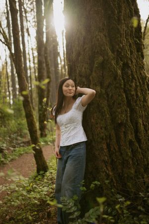 Person in a white top and jeans poses against a large mossy tree trunk in a misty forest setting.