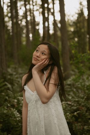 Person in a white lace dress poses dreamily in a serene forest setting during golden hour.