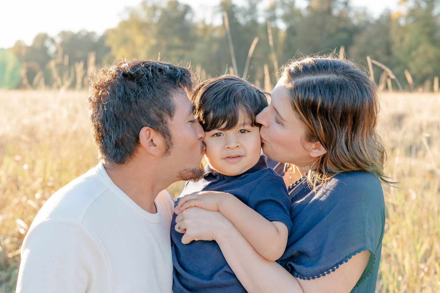 Loving parents kiss their young dark-haired toddler on the cheeks in a sunny field.