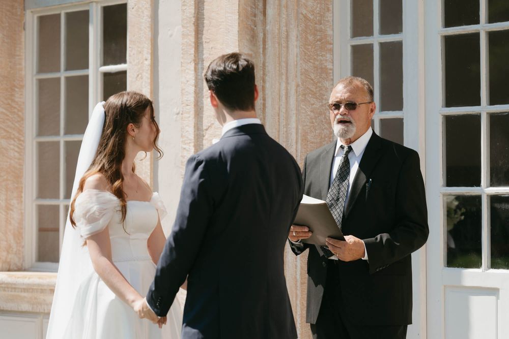 A wedding ceremony takes place outside a stately stone building with large windows.
