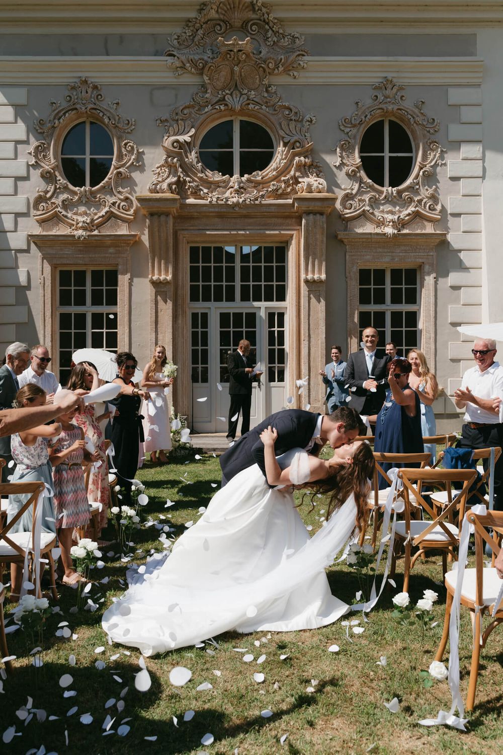 A dramatic wedding dip kiss surrounded by flower petals and guests outside an ornate building.