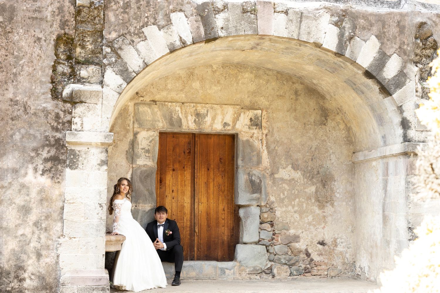 Romantic wedding portrait under rustic stone archway with wooden door.