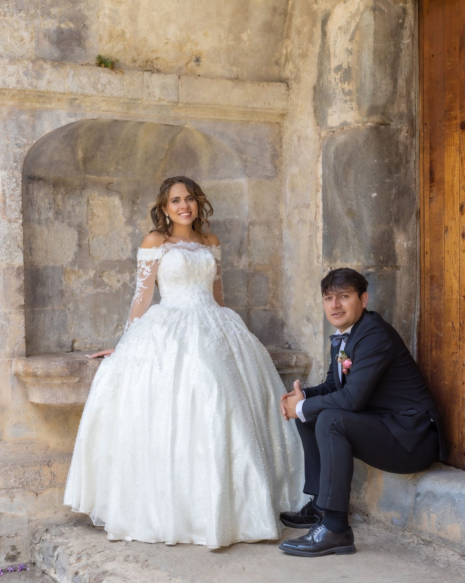 Wedding couple in formal attire posing against stone wall architecture.