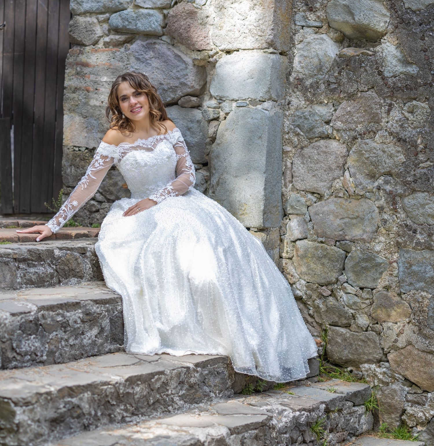 Bride in luxurious lace gown seated on stone steps against rustic wall.