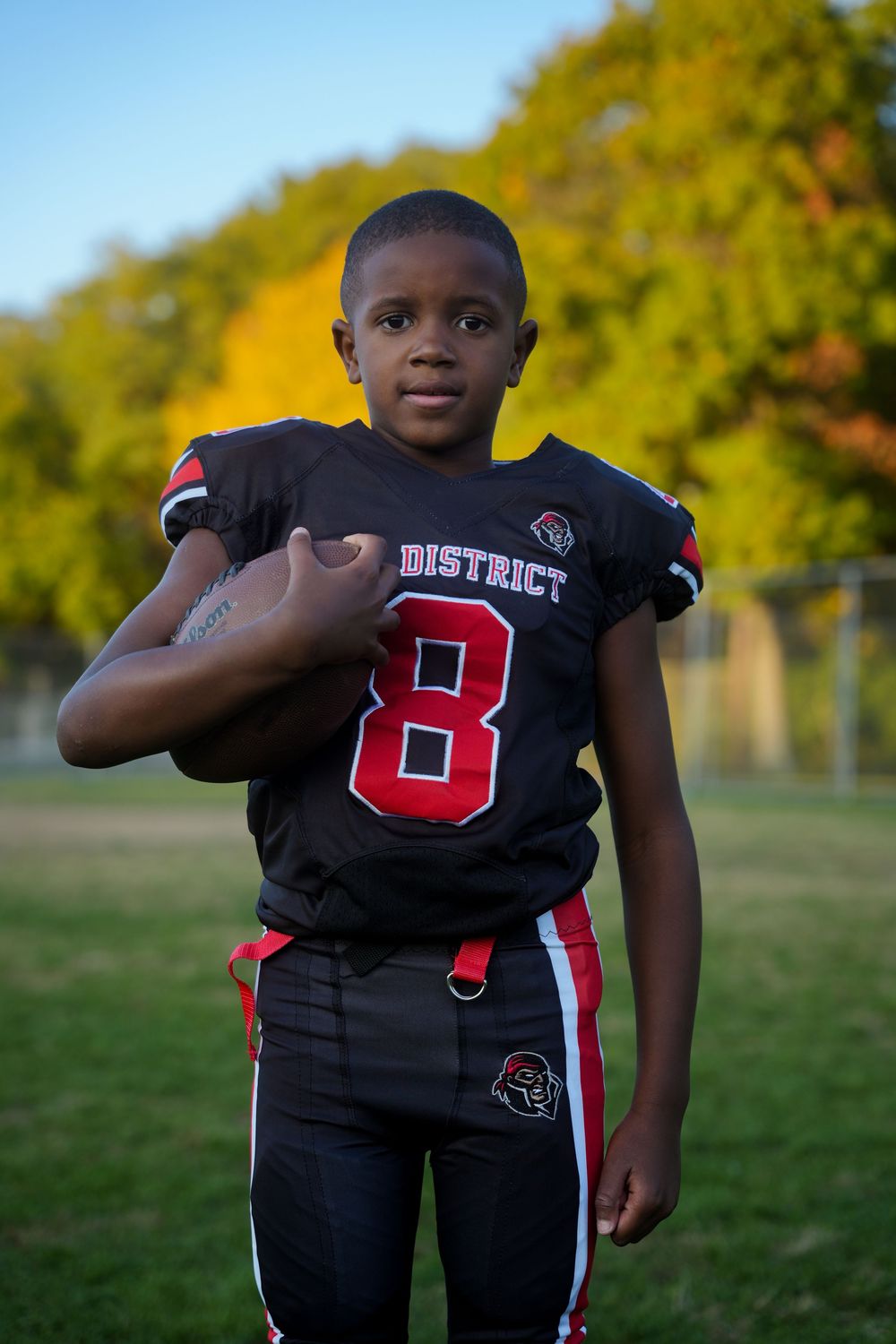 Young football player in black and red uniform number 8 poses on field during golden hour.