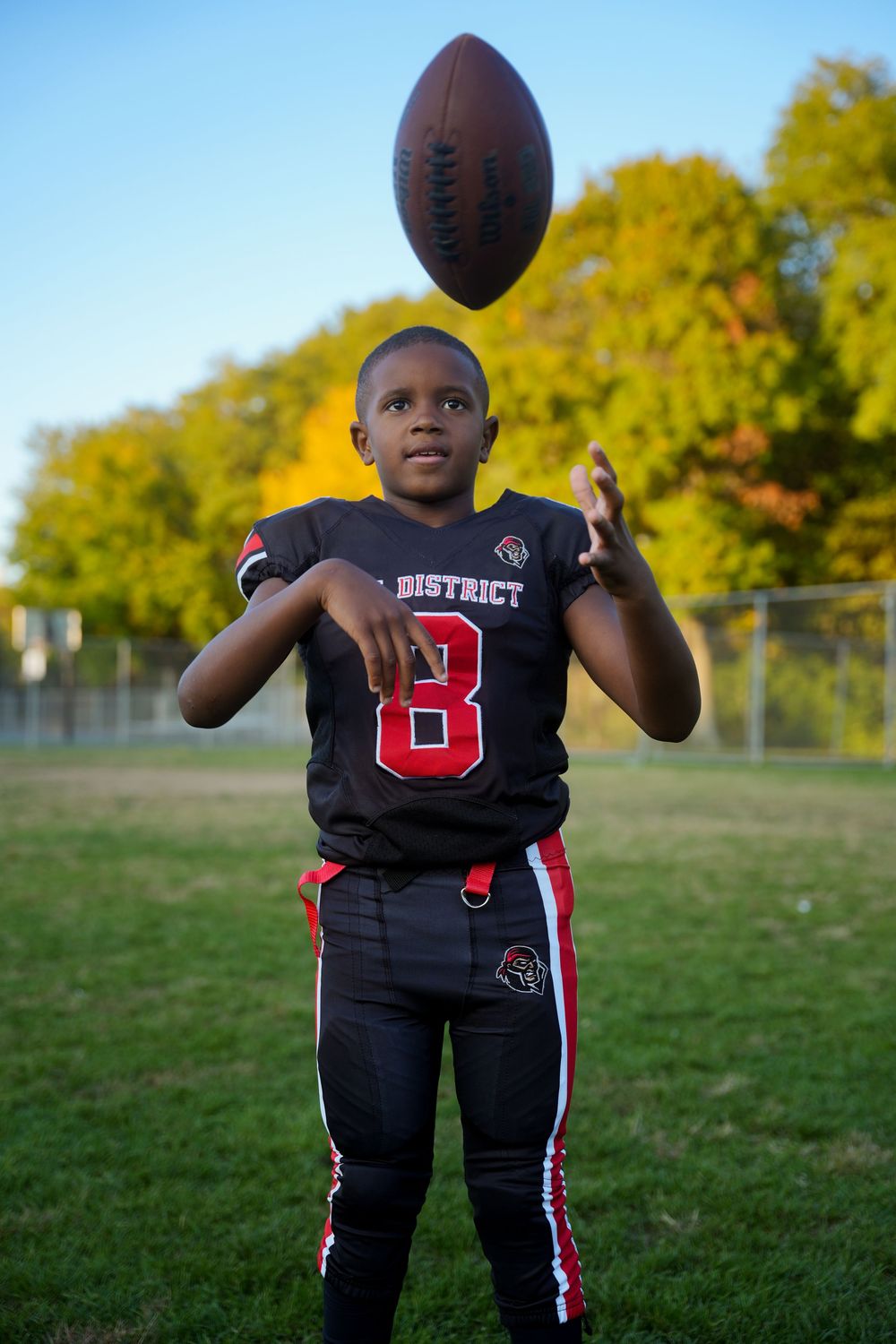 A football player in a black and red uniform poses with a football on a grassy field during autumn.