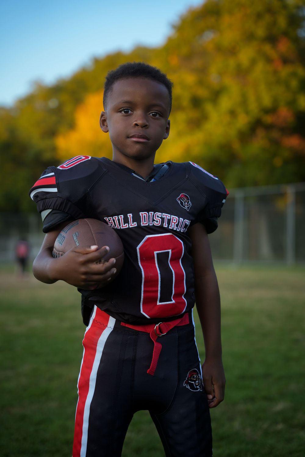 A young football player in a black and red uniform practices on a field during autumn with golden trees in background.
