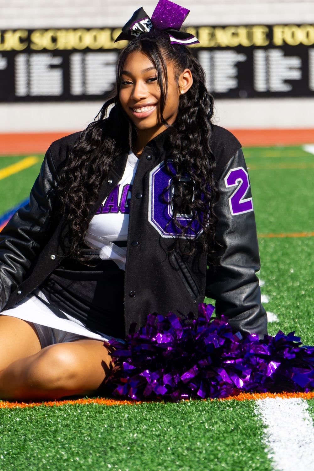 Cheerleader in purple and black uniform poses on artificial turf field with pom poms during photo session.