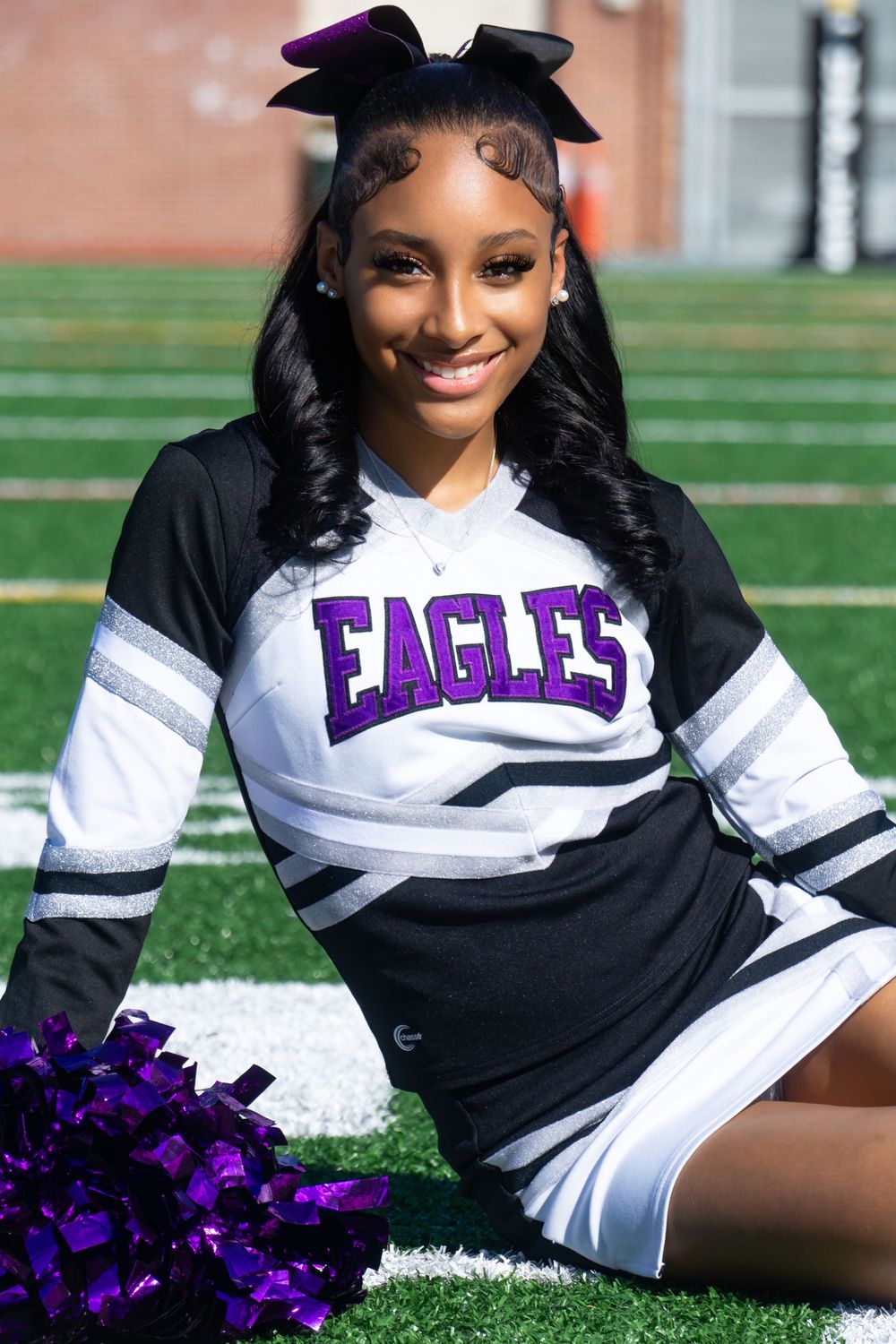Cheerleader in black and white uniform with purple Eagles text poses with pom poms on football field.