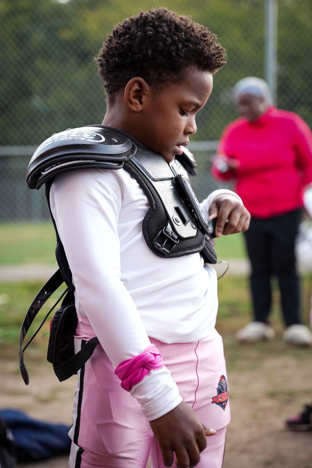 A young child in a pink and white outfit carries a black football helmet while walking outdoors on a field.