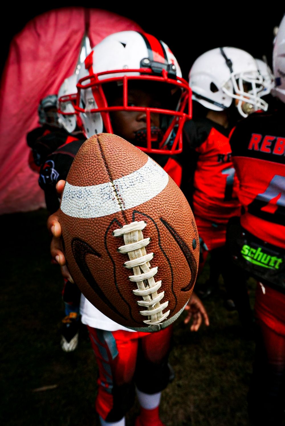 Close-up sequence of a brown leather football being held against a backdrop of red sports equipment and helmets.