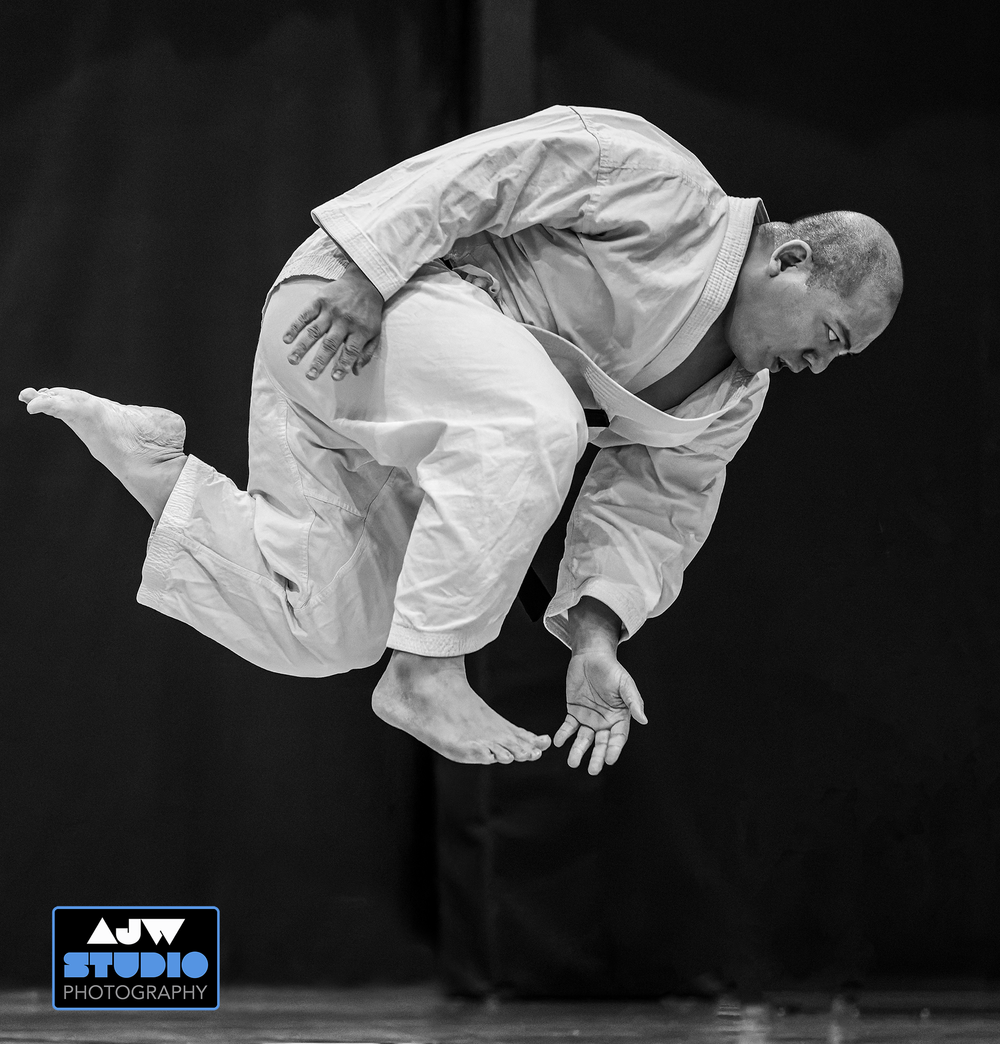 A martial artist in white gi executes an acrobatic jump technique against a black background.