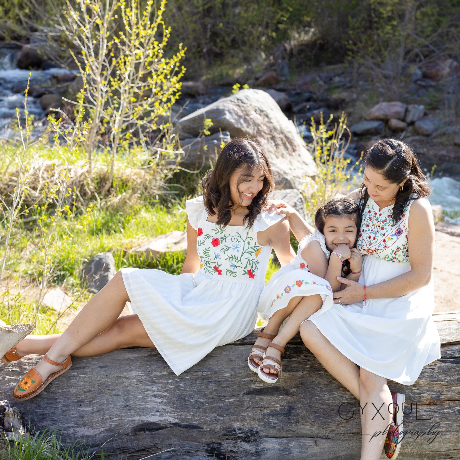 Three people in white dresses sit together on rock by rushing river.