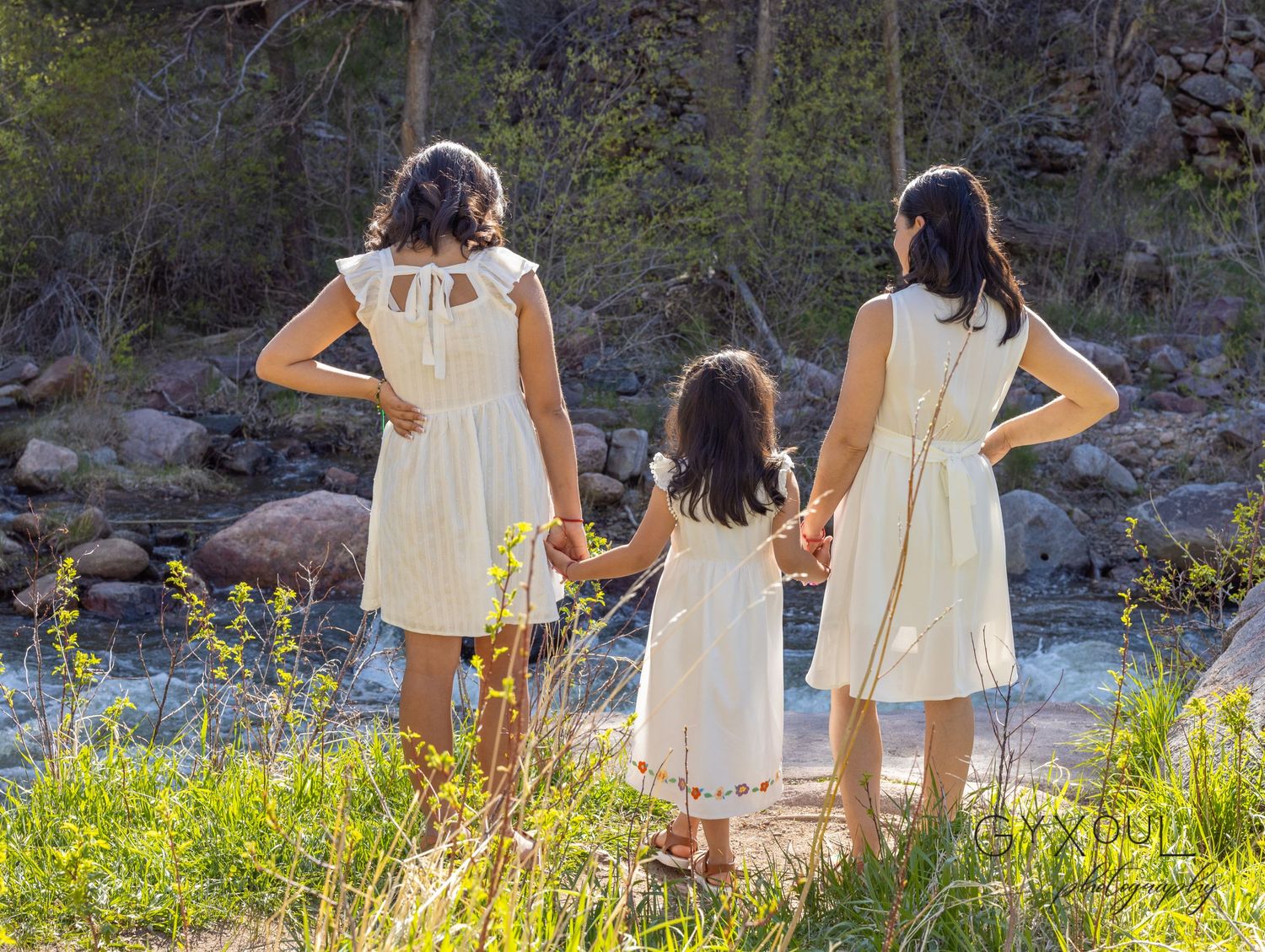 Three people in matching white dresses stand looking at river scenery.