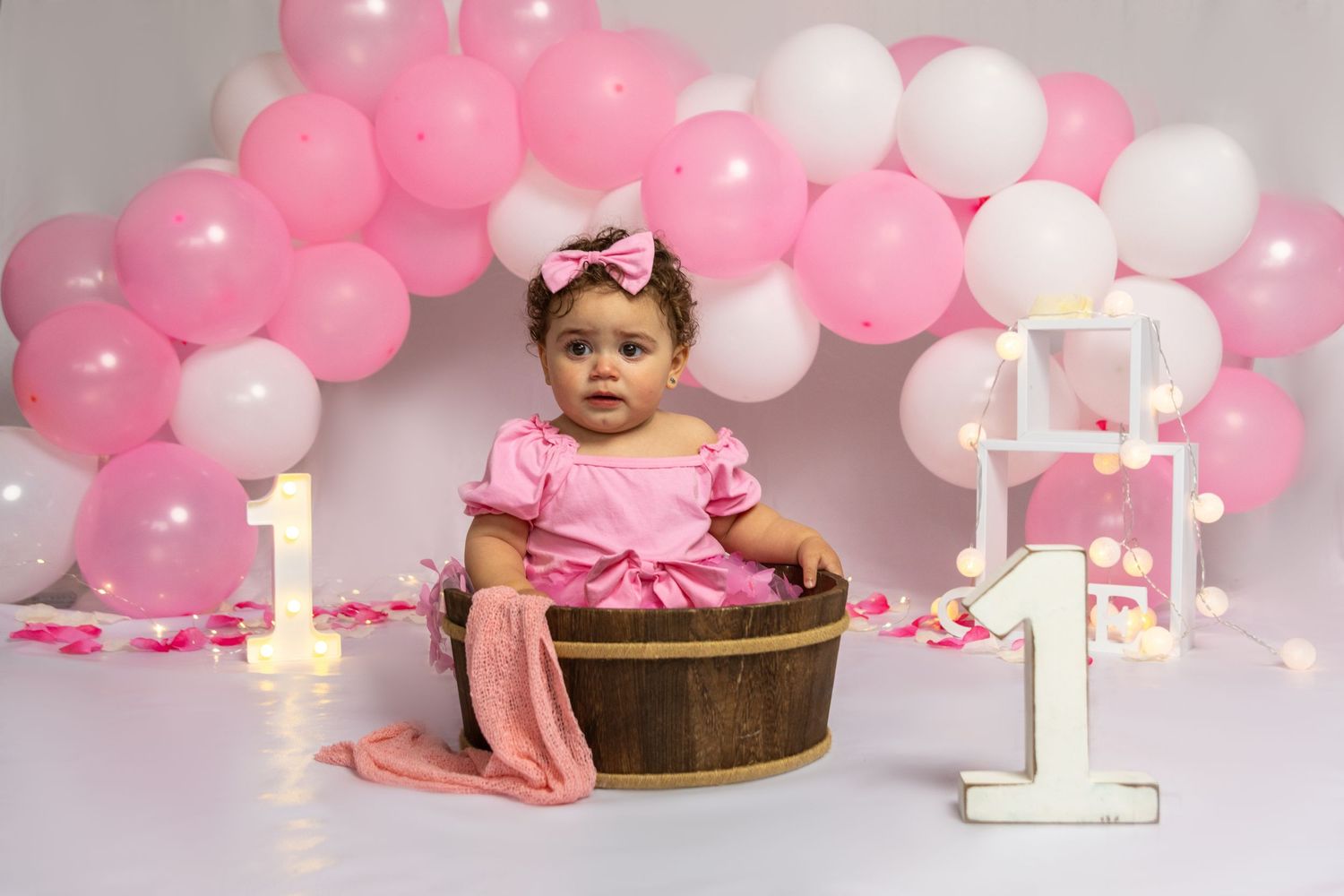 Pink and white balloon arch with wooden stool and illuminated number one for birthday photos.