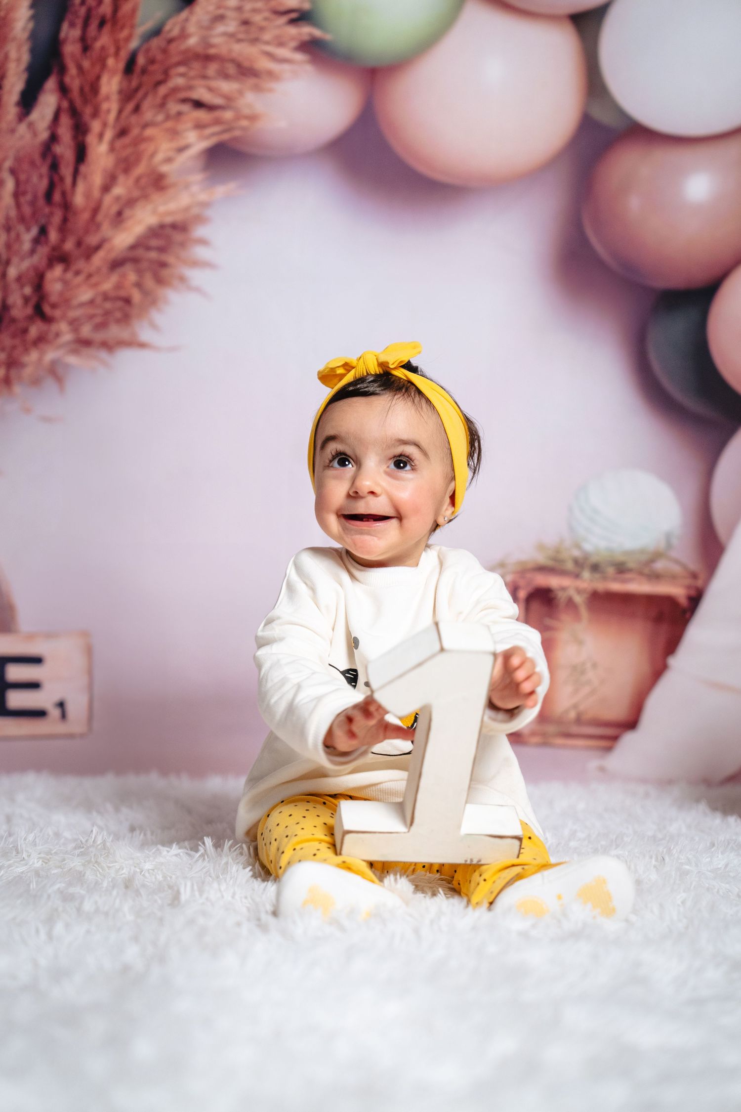 First birthday portrait with yellow accessories and number one prop against pink balloon backdrop.