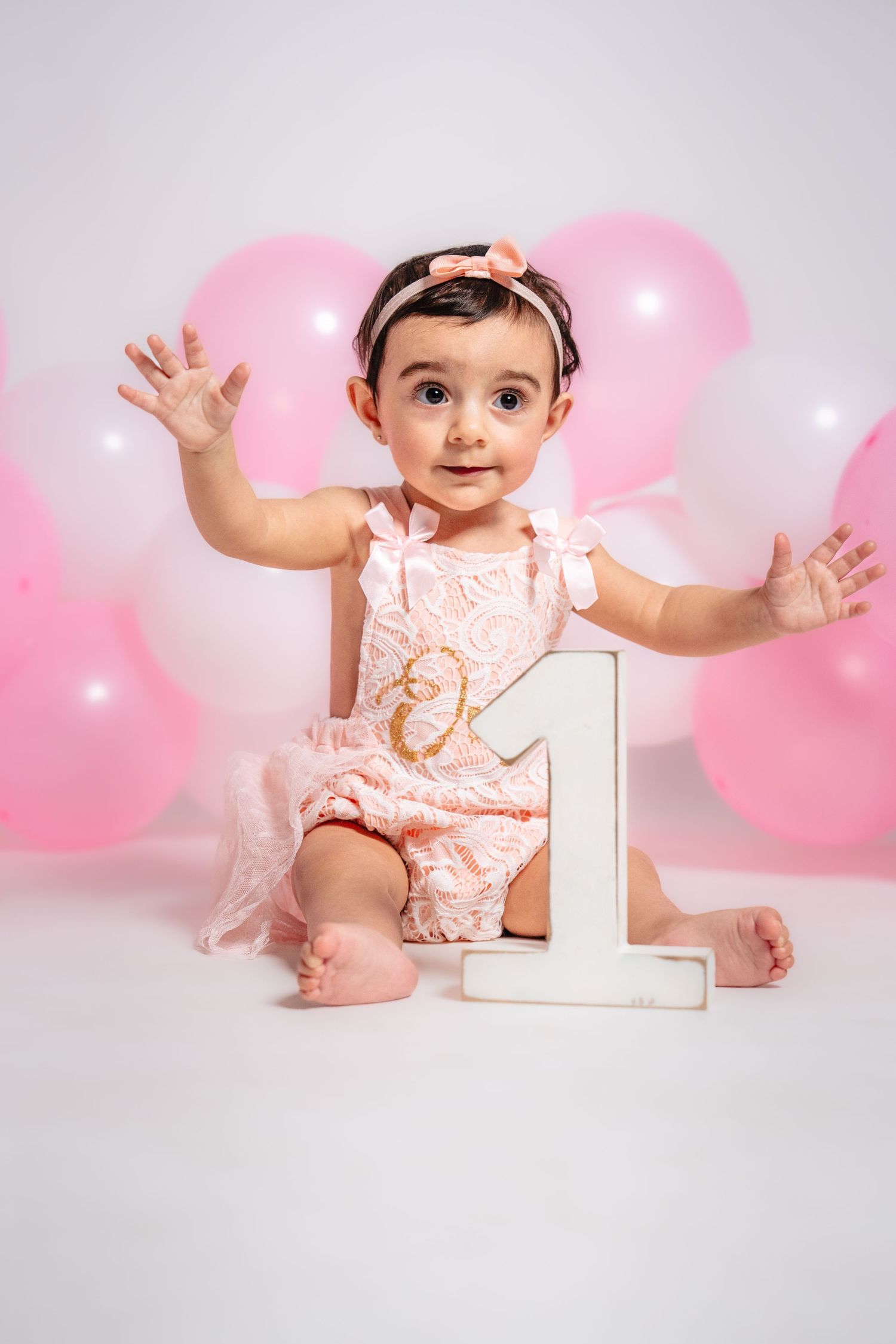First birthday photo with pink balloons and number one prop in soft lighting.