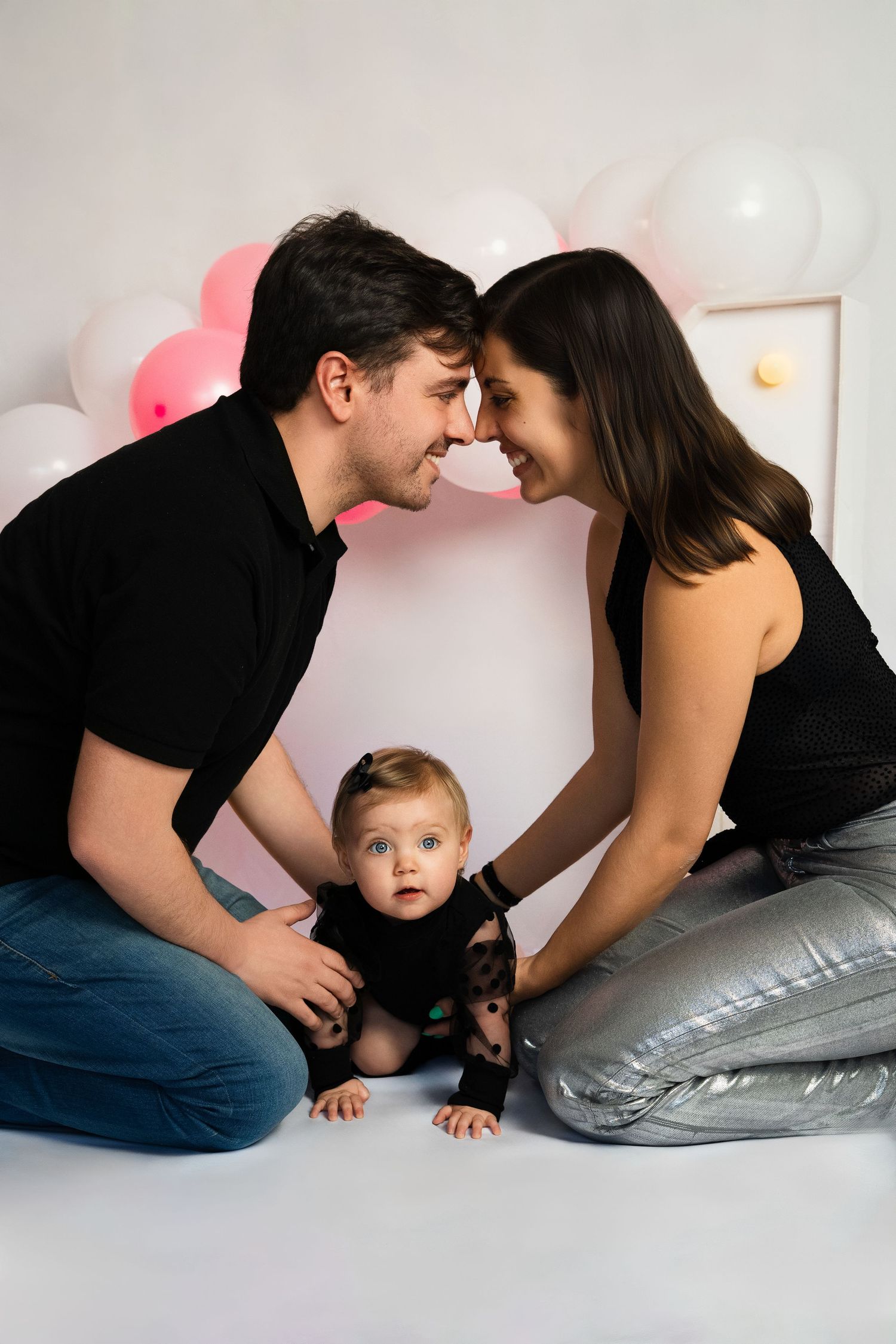 Family first birthday portrait with pink balloon arch and string lights in background.