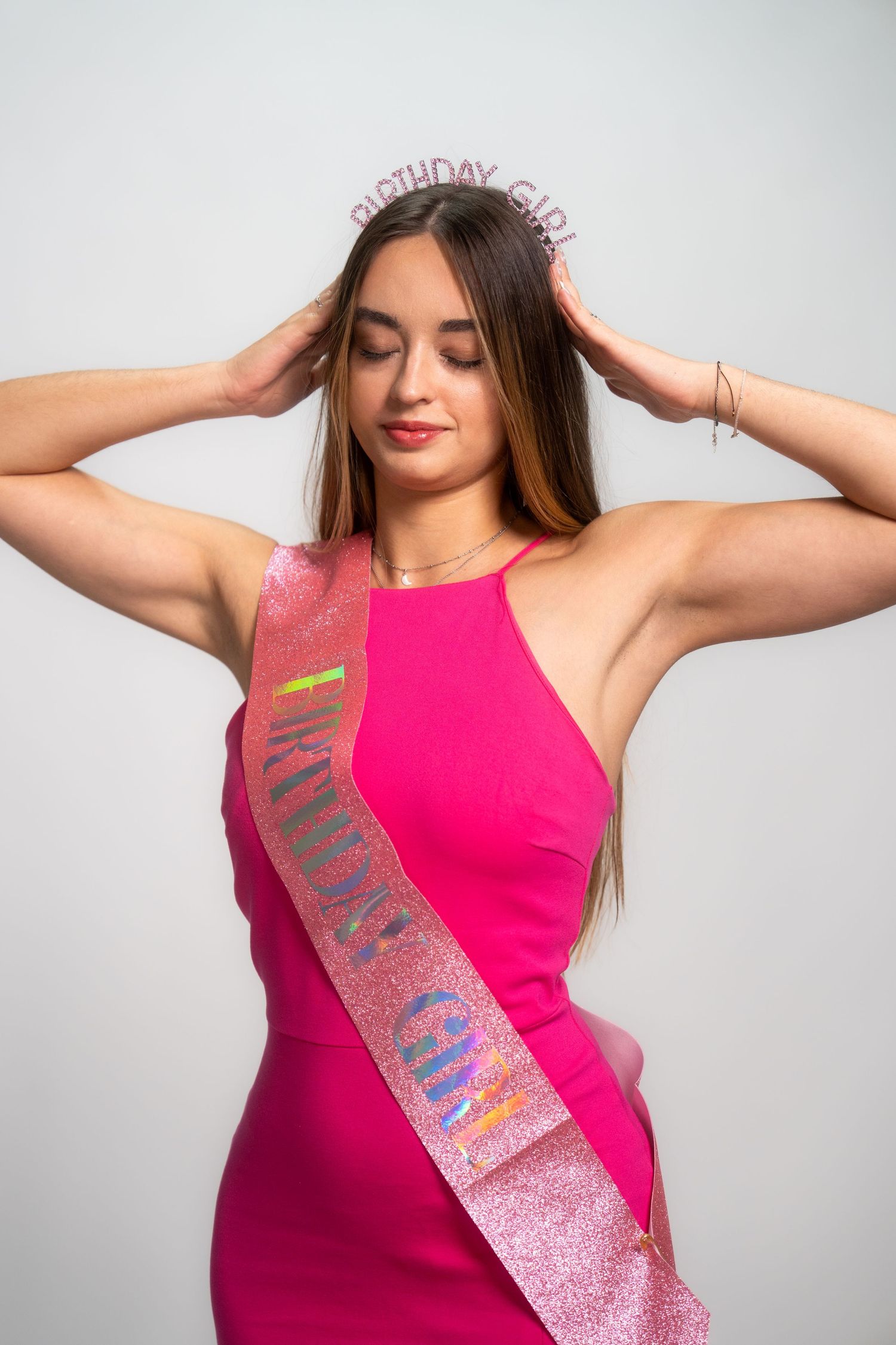 Model wearing pink dress and celebration sash poses against white background for birthday photos.