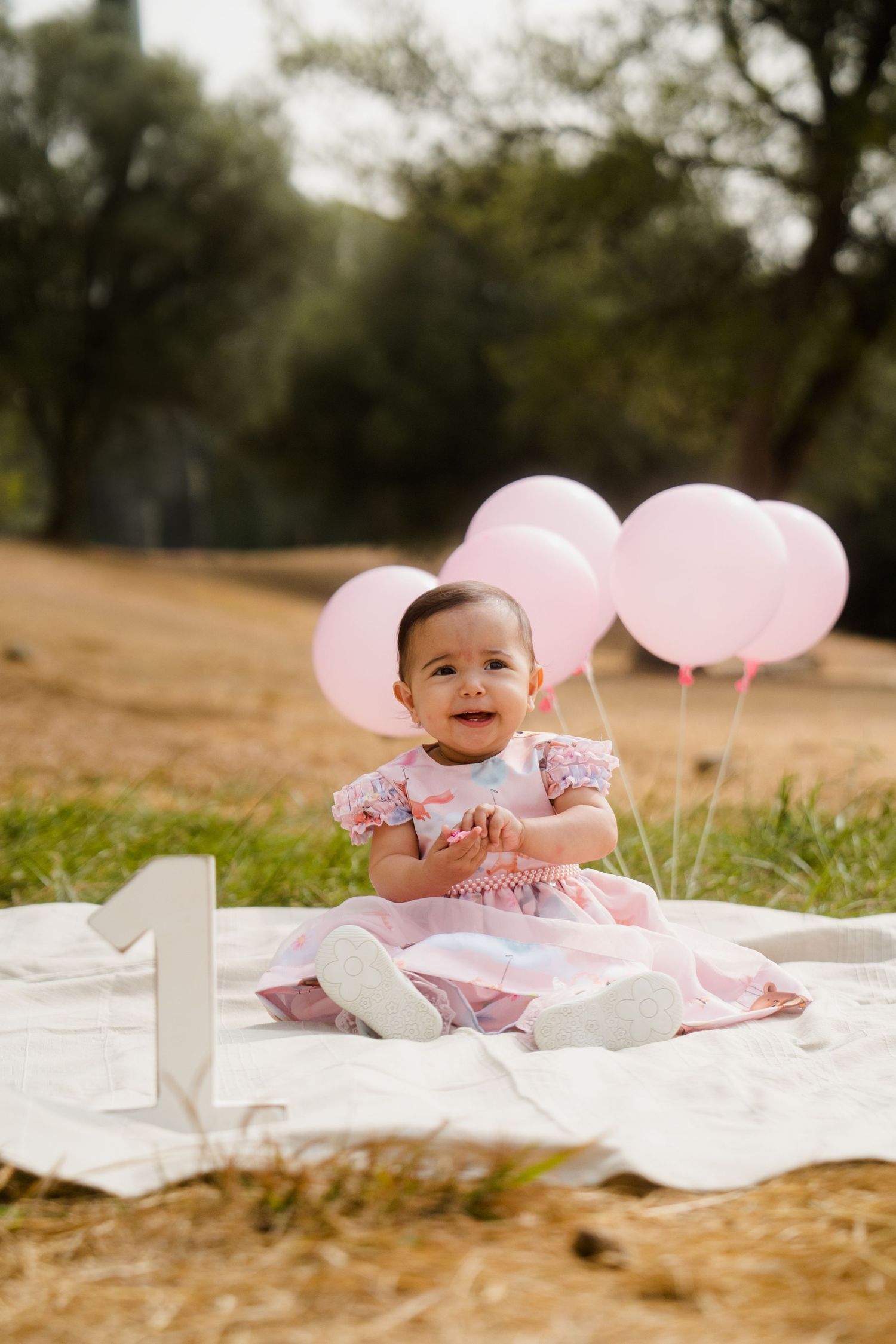 Baby photoshoot on white blanket with pink balloons in outdoor setting.