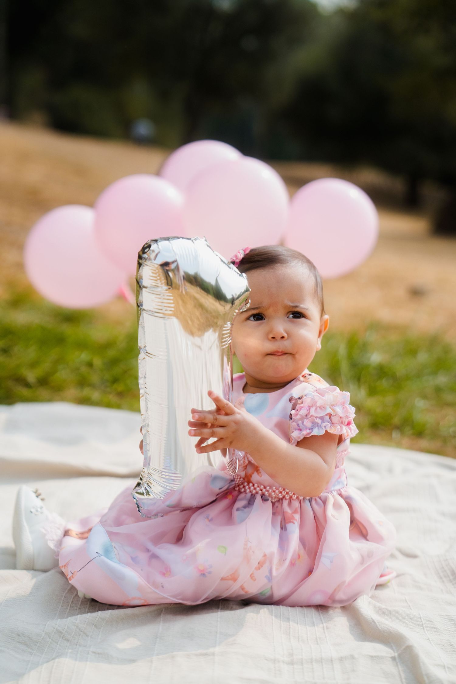 A pink dress adorned baby lying on a white blanket in an outdoor setting.