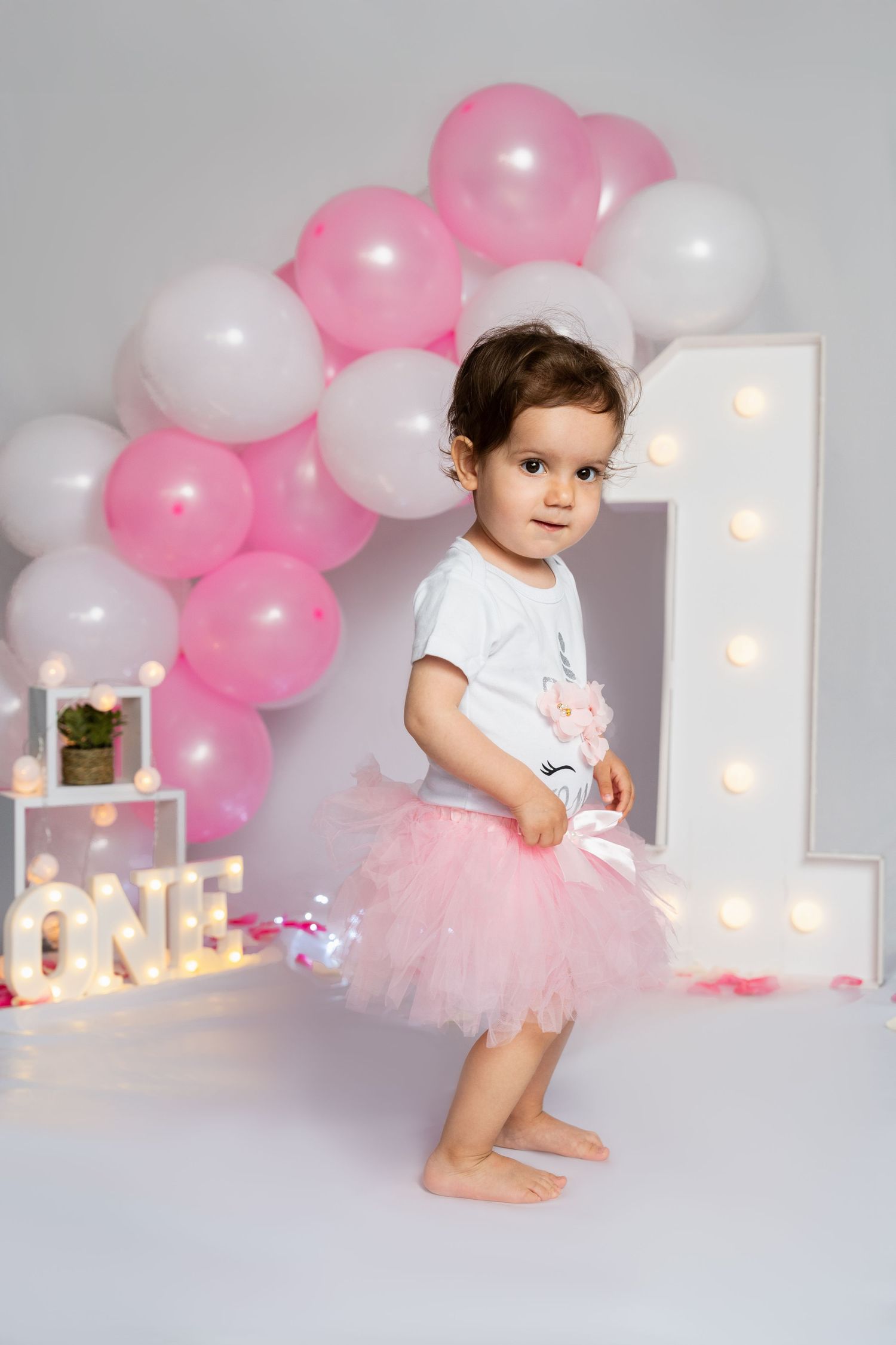 First birthday setup with pink and white balloons, illuminated number one, and tutu outfit.