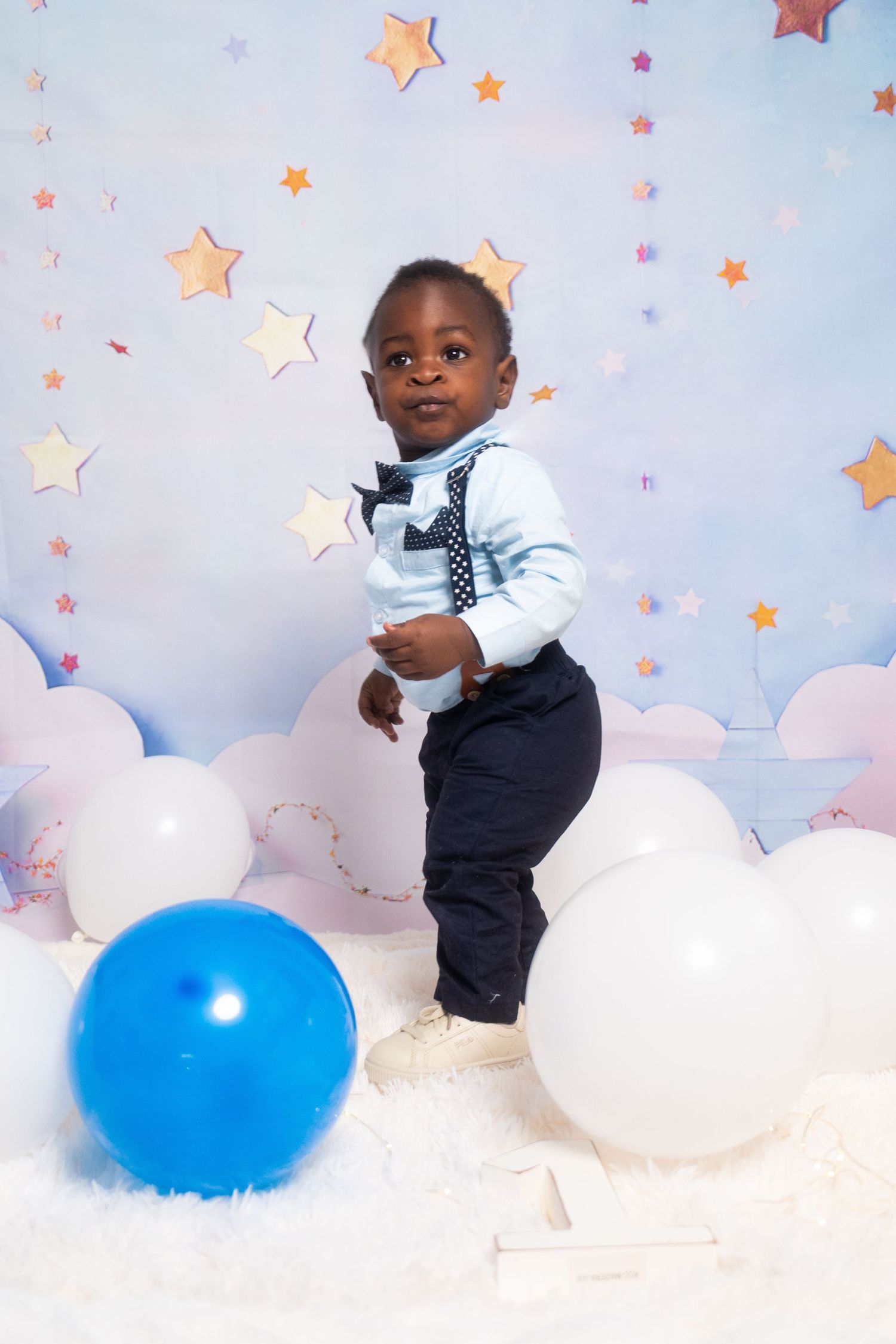 Baby in navy outfit playing with blue and white balloons against starry backdrop.